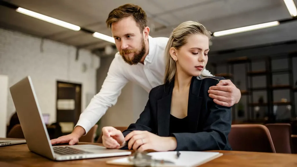 A man leaning over a woman's shoulder at her laptop in a dim office