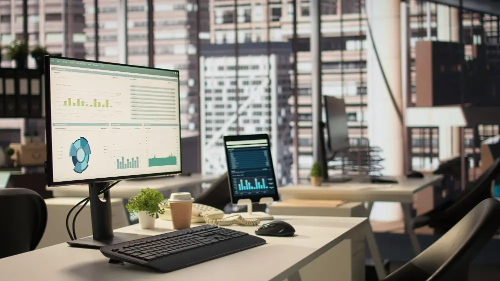 Empty office desk with an operations dashboard on the monitor and a tablet showing performance charts