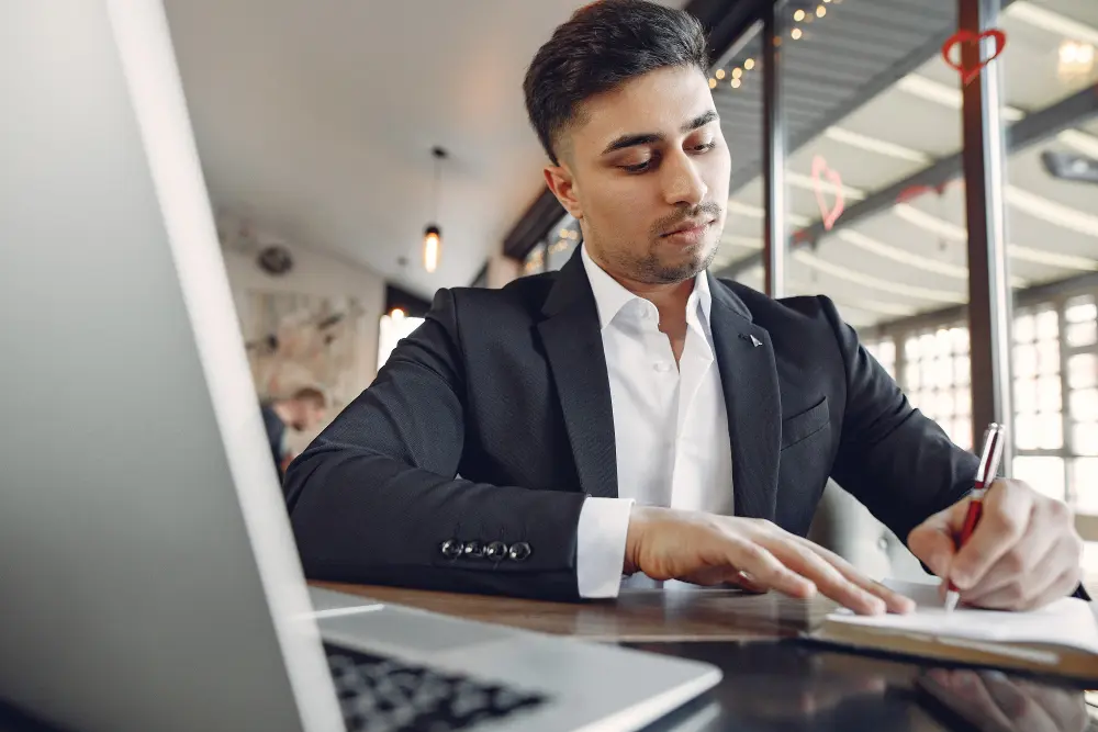 A man in a suit taking notes beside a laptop